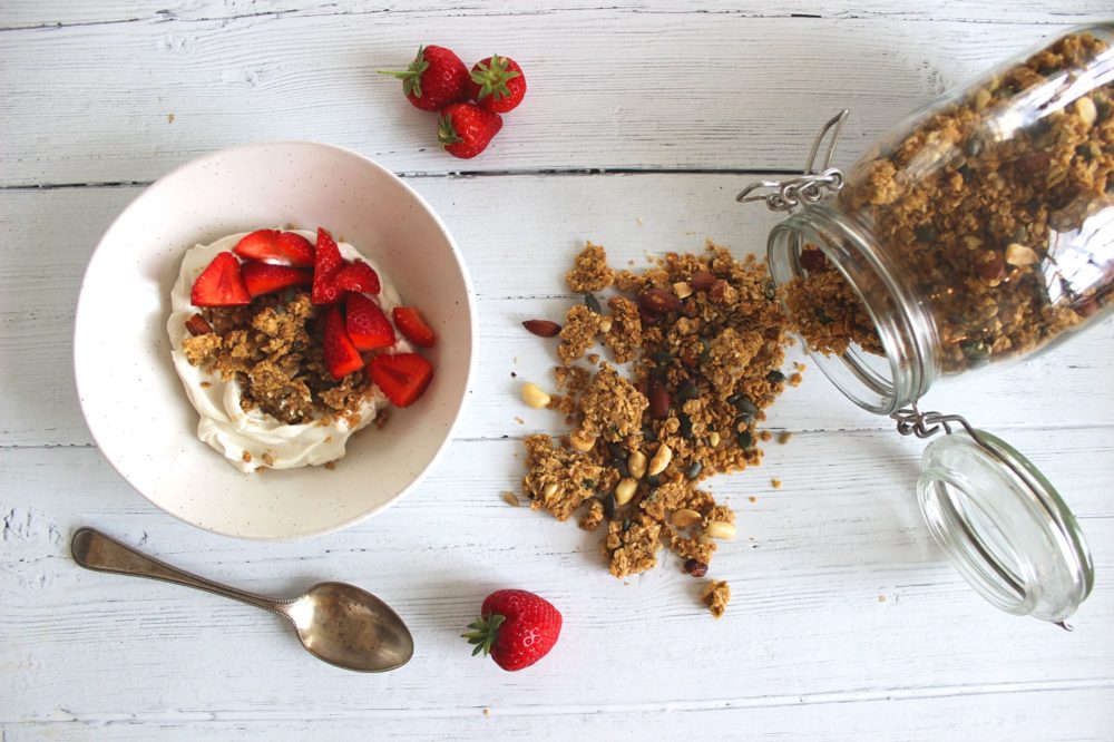 peanut butter granola coming out of a clear jar next to a bowl with strawberries against a white background