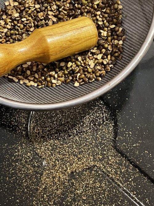 mung beans and wooden pestle in a sifter against a black countertop.