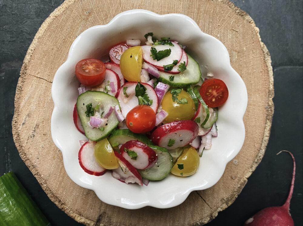 cucumber and radish salad with red onions and tomatoes in a white scalloped bowl on a black countertop.