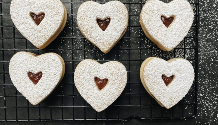 heart shaped vegan linzer cookies with red jam visible, dusted with powder sugar and cooling on a wire rack against a black countertop background.