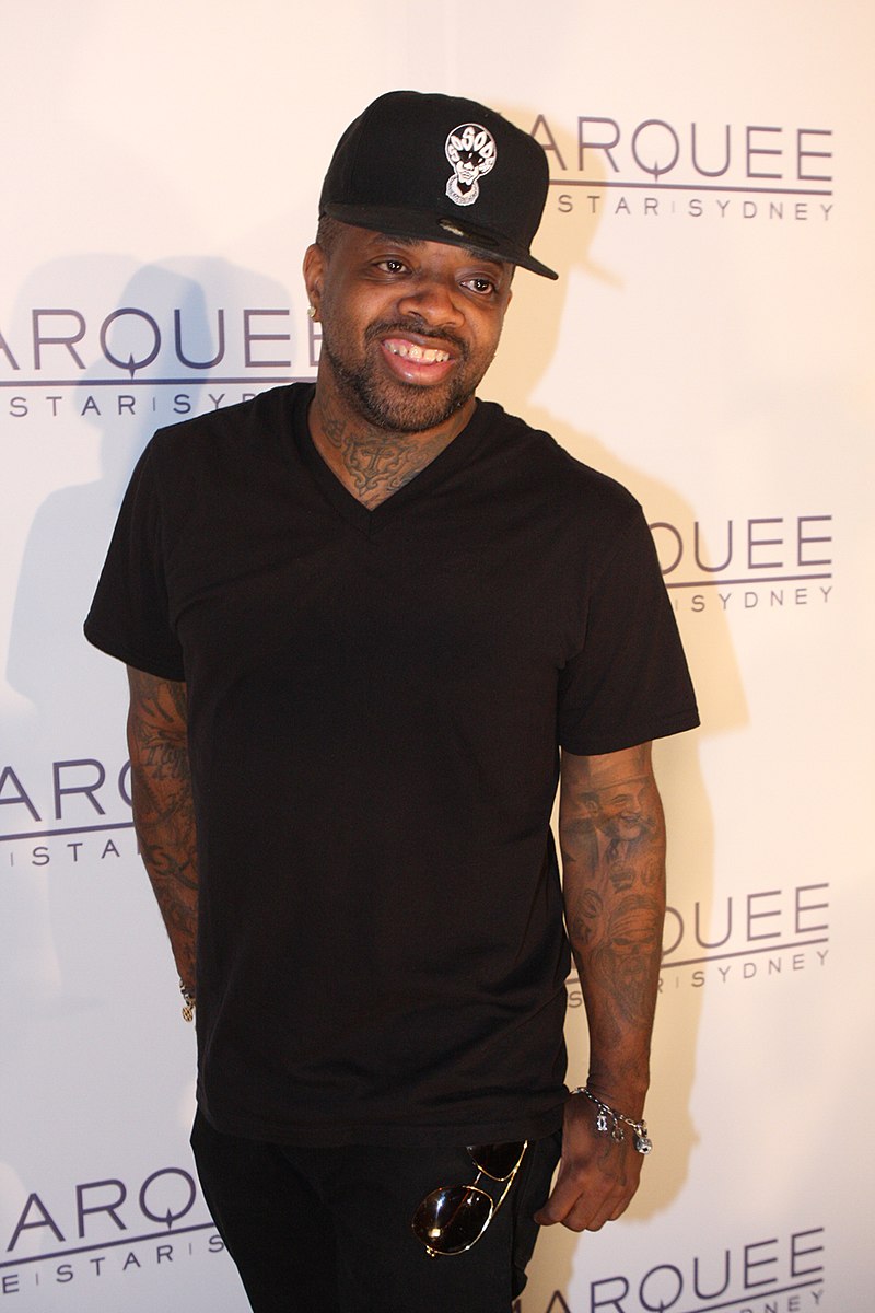 Jermaine Dupri wearing black trucker hat, black t-shirt, and black pants against a step and repeat background.